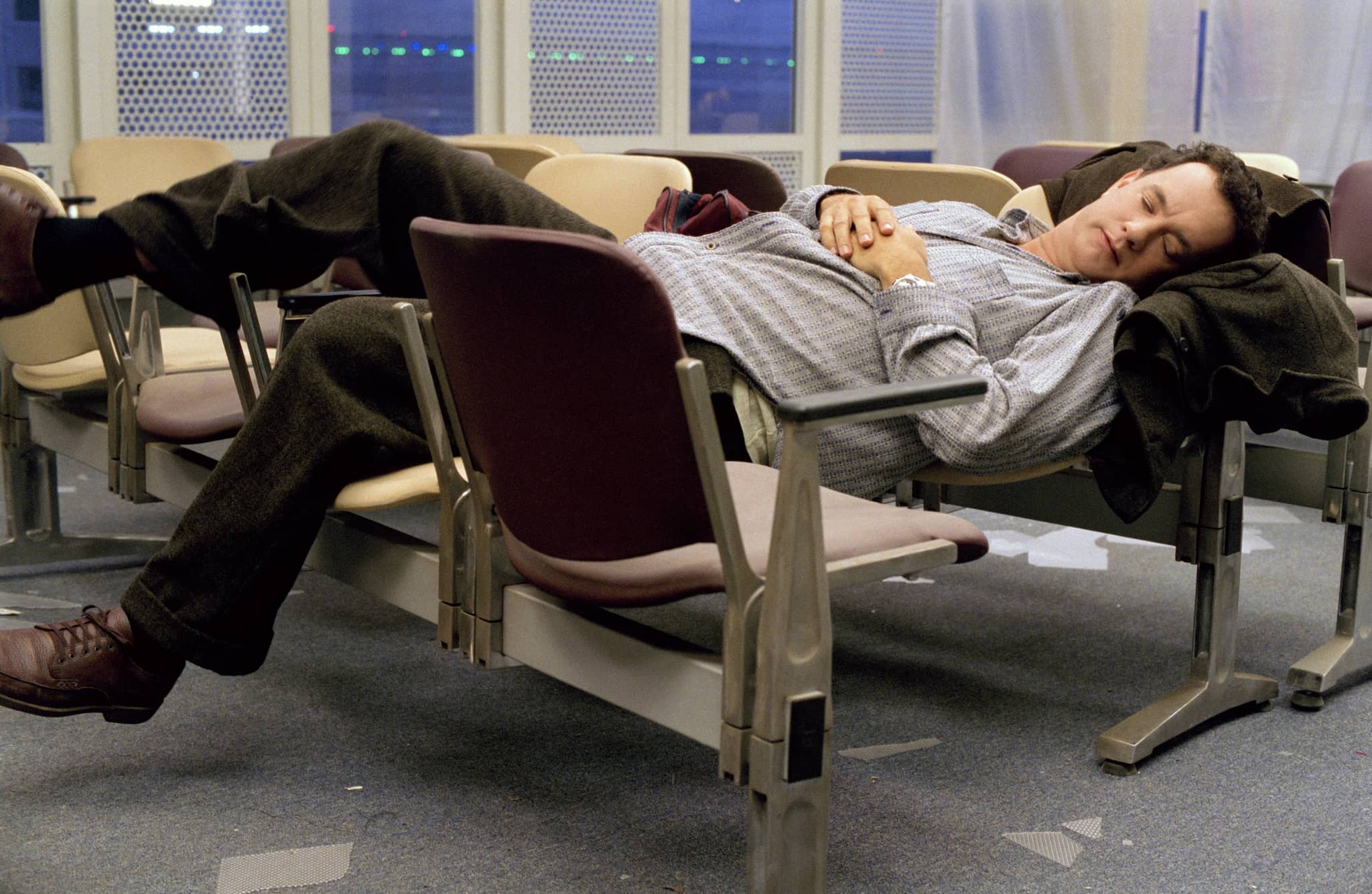 A middle-aged man uncomfortably sleeping on airport chairs with a coat under his head.